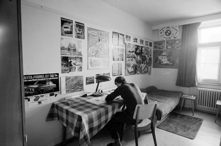 Photo en noir et blanc d'une chambre dans l'institution éducative Uitikon-Waldegg. Un adolescent est assis à son bureau de travail, à gauche se trouve un lit avec un tapis devant. Des rideaux ornent la fenêtre et des posters de voitures de sport décorent les murs.