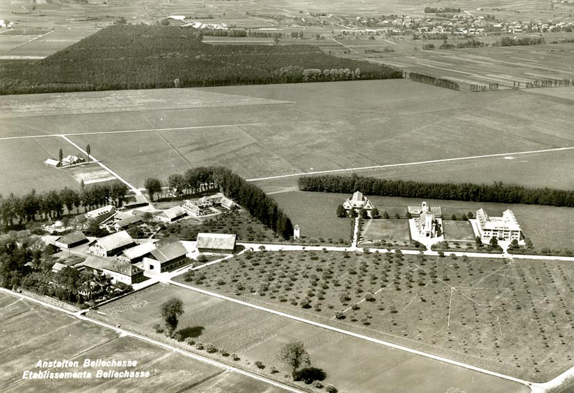 La photographie aérienne montre à gauche les baraques d'habitation des internés ainsi que les ateliers et les bâtiments économiques, entourés d'arbres. À droite se trouvent la villa du directeur, l'église et l'aile de la prison