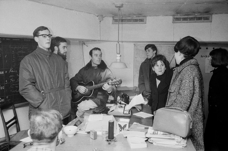 Photo en noir et blanc du jeune pasteur Ernst Sieber au centre, tenant une guitare, dans une pièce de bunker. Autour d'une table, six autres personnes se tiennent debout ou sont assises, toutes vêtues de vêtements d'hiver. Elles écoutent la musique ou discutent.