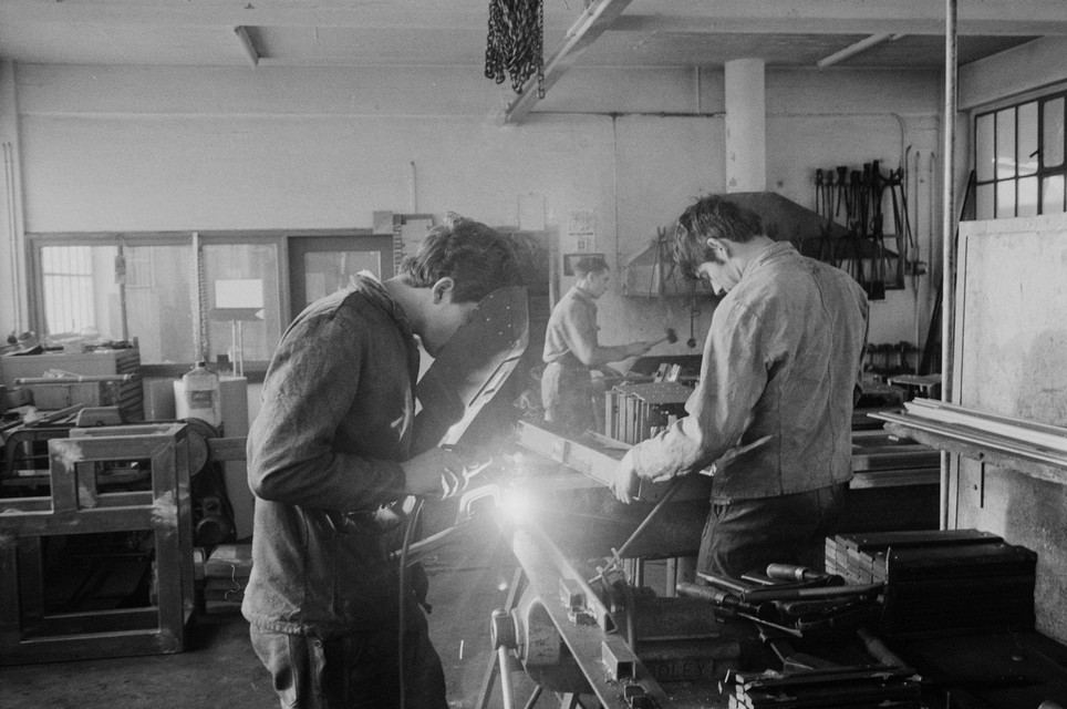 Trois apprentis travaillent dans un atelier de serrurerie. Au premier plan, on soude, et à l'arrière, le métal est travaillé avec des outils.